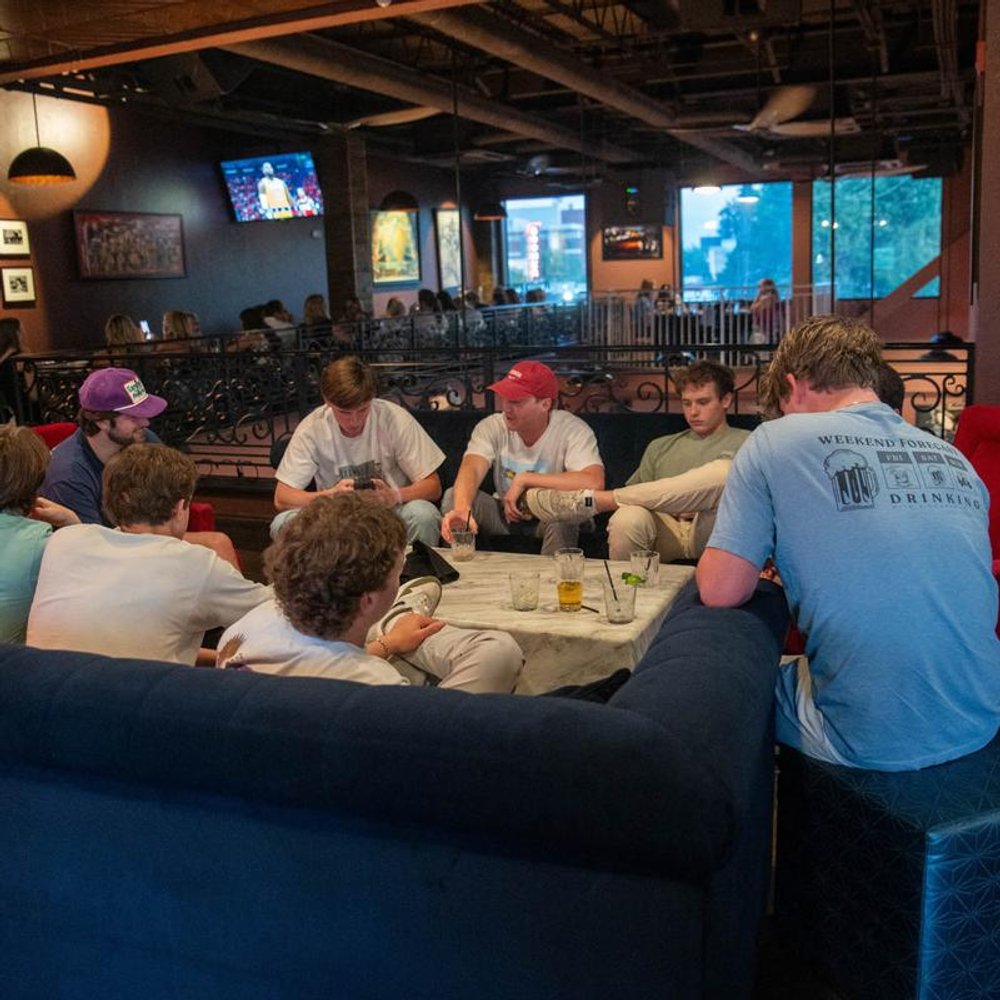 Group Of People Socializing Around A Table With Drinks In A Bar Setting.