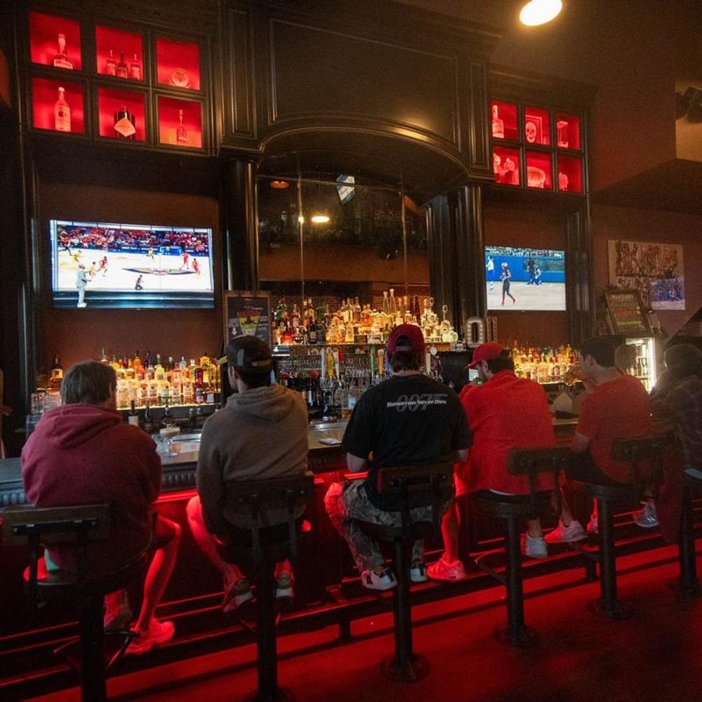 Guests At A Bar Facing Away With Tvs And Liquor Bottles On Shelves.