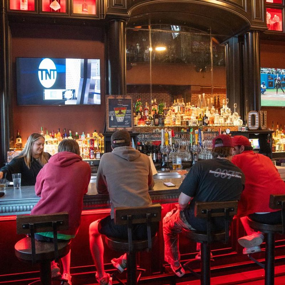 Guests Seated At A Bar Facing Away Liquor Bottles On The Shelves Tvs.