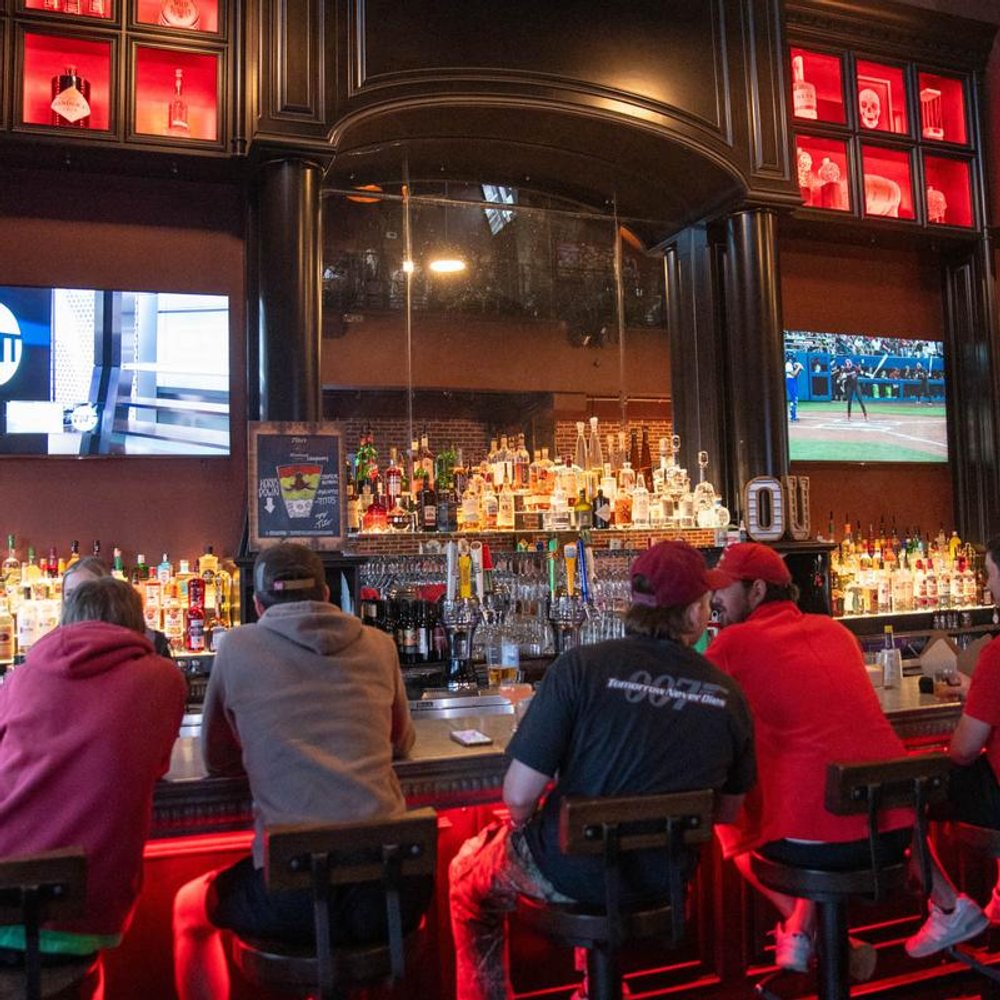 Guests Sitting At A Bar With A Variety Of Liquor Bottles And Tvs Playing Sports.