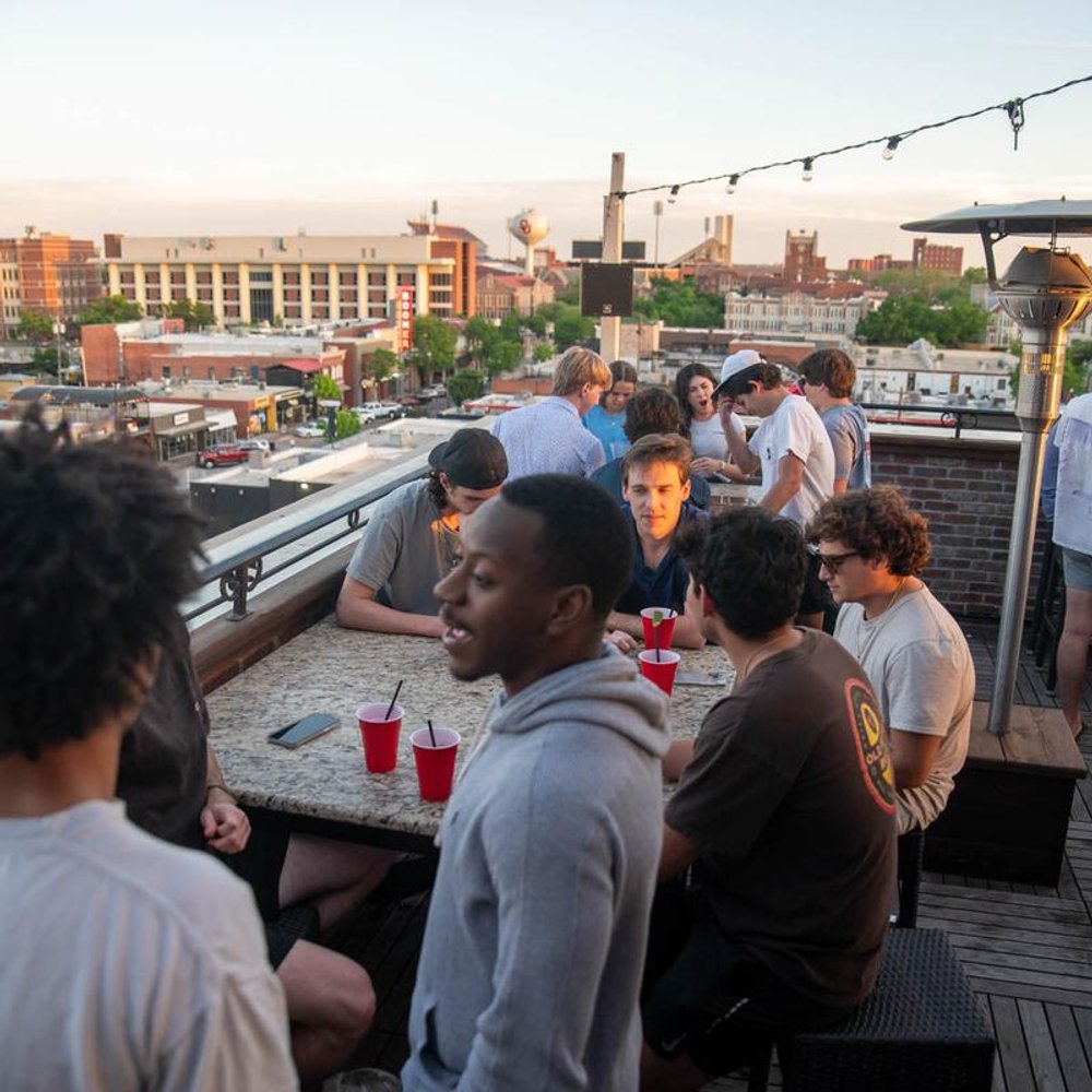 People Socializing On A Rooftop Patio With Cityscape In The Background.
