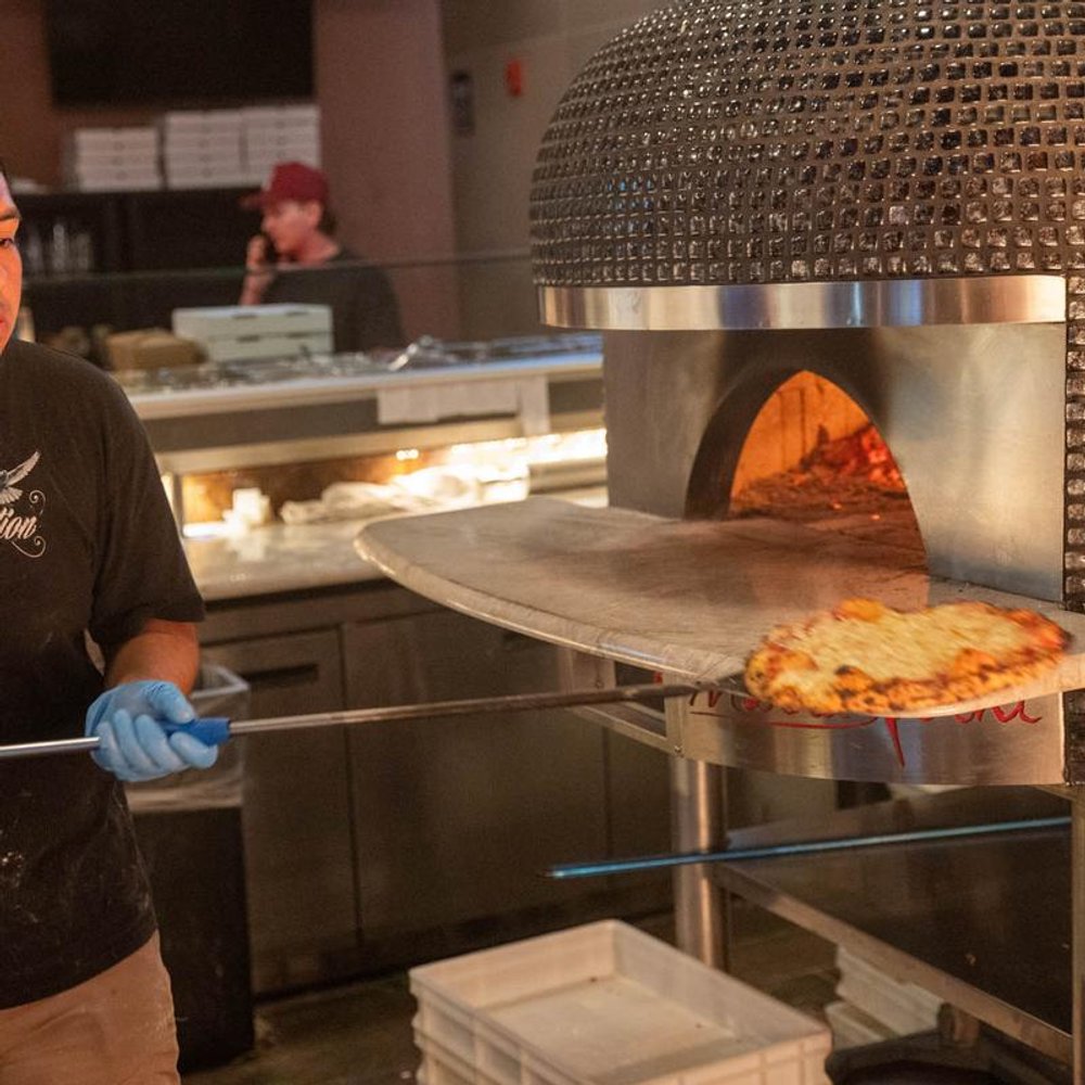Person In Gloves Removing A Pizza From The Wood Fired Oven With A Peel.