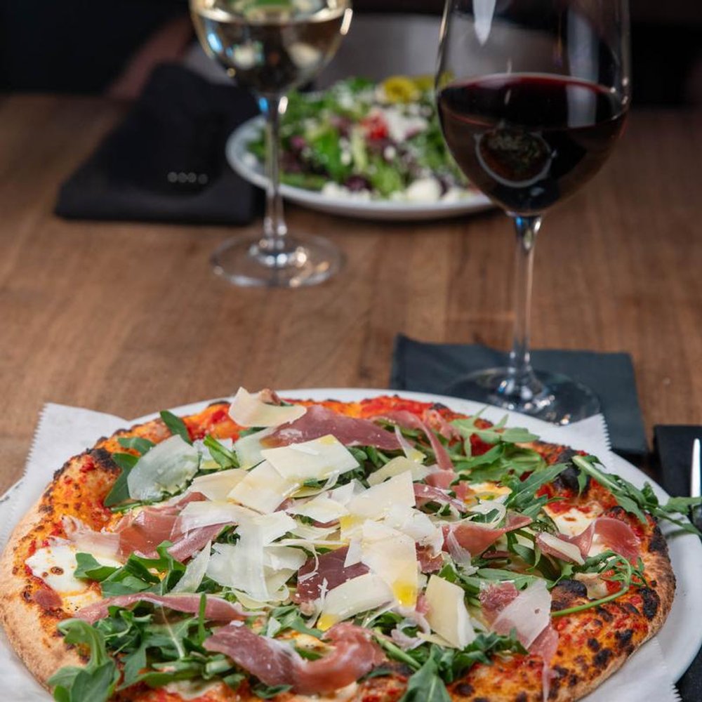 Prosciutto Arugula Pizza With Parmesan On A Table With Glasses Of Wine And A Salad.