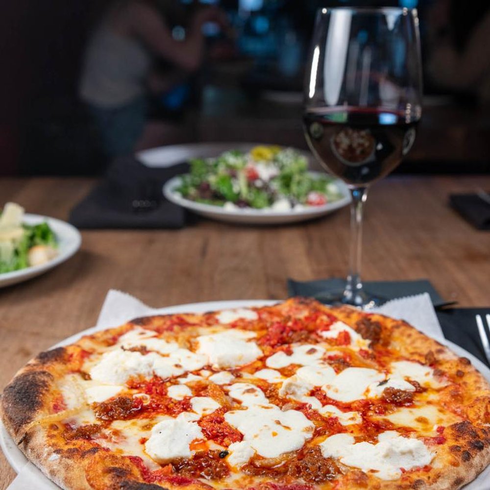 Red Pizza With Cheese On A Table With Red Wine Salads In The Background.